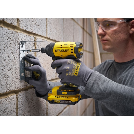 Person uses Stanley V20 cordless impact driver on masonry wall while wearing gloves and safety glasses.