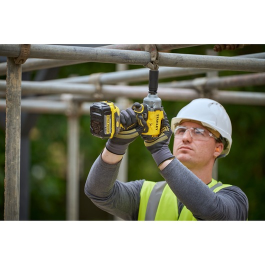 Worker uses STANLEY SFMCF921B cordless impact wrench on scaffolding, wearing safety gear and gloves.