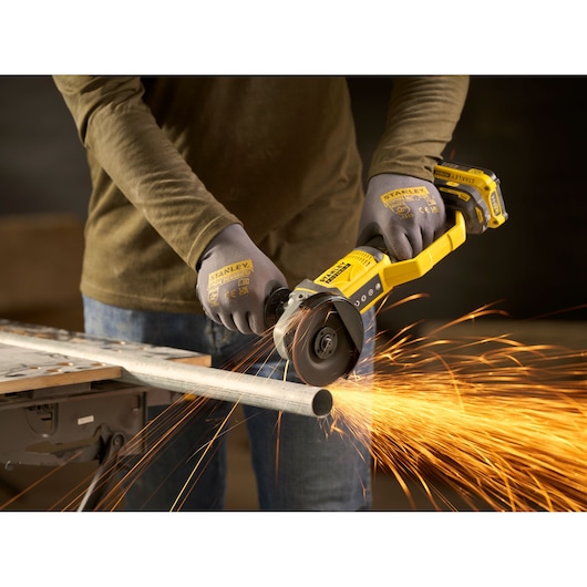 Photo of STANLEY SFMCG300B cordless angle grinder cutting metal pipe, sparks flying, worker wearing gloves.