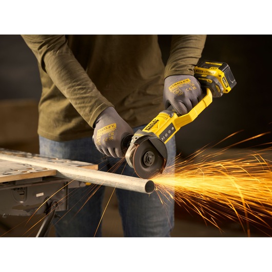 Photo of STANLEY SFMCG300B cordless angle grinder in use cutting metal pipe, producing bright sparks.