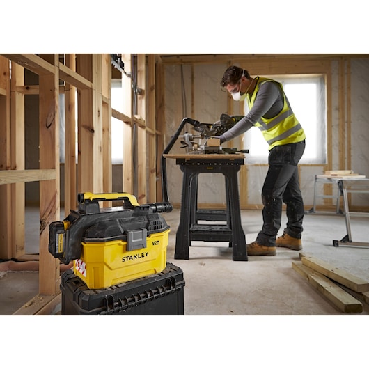 STANLEY V20 vacuum cleaner on a toolbox at a construction site, worker operating saw in the background.