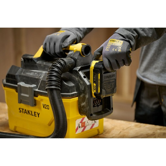Stanley V20 cordless vacuum in use, person with gloves installing battery on yellow and black unit.