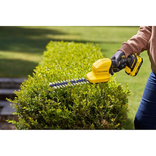 STANLEY STCSS20D1 yellow cordless hedge trimmer in use trimming green bushes outdoors.