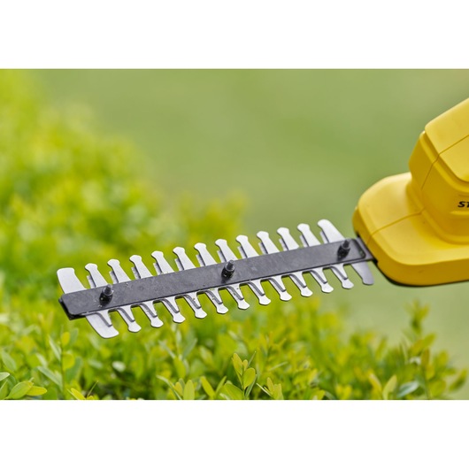 STANLEY STCSS20D1 hedge trimmer blade close-up above green bushes, side angle, yellow casing visible.