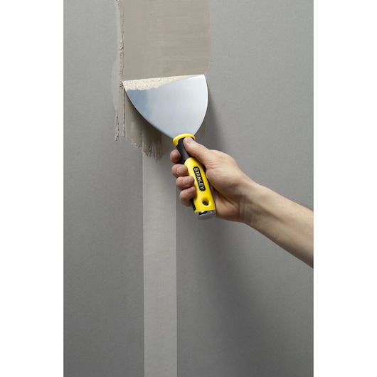 Photo of STANLEY STHT0-05799 putty knife with yellow handle applying filler to wall, close-up.