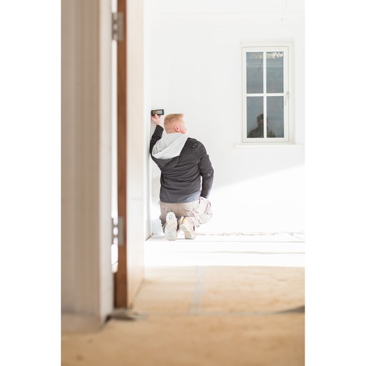 STANLEY STHT1-77139 laser distance measurer in use by a person kneeling near a white wall in a bright room.