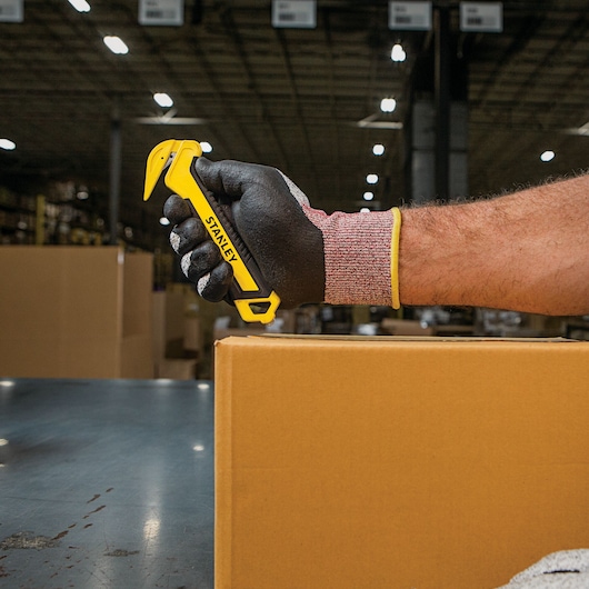 STANLEY STHT10356 safety box cutter held by a gloved hand, yellow handle, warehouse setting.