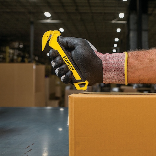 Photo of STANLEY STHT10356 yellow safety knife held in gloved hand, cutting cardboard box in warehouse.
