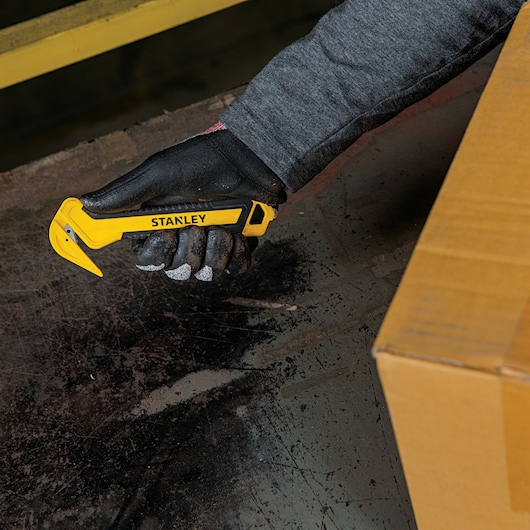 Stanley STHT10356 utility knife held in gloved hand near cardboard box, yellow and black design.