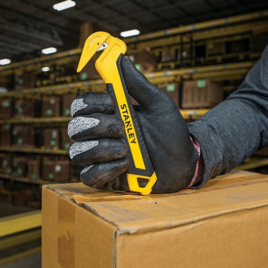 Stanley STHT10356 safety box cutter held by a gloved hand opening a cardboard box in a warehouse.
