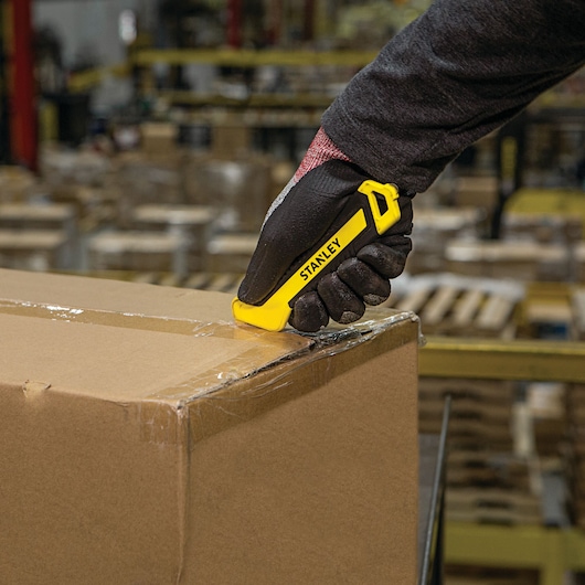 STANLEY STHT10356 yellow utility knife cutting tape on a cardboard box, warehouse background, product in use.