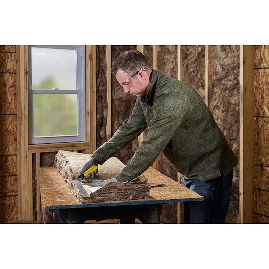 A person wearing gloves uses a yellow and black STANLEY utility knife to cut insulation material on a workbench in a room under construction.
