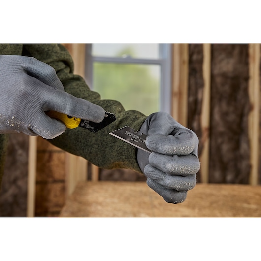Close-up of a person wearing grey work gloves, inserting a STANLEY utility knife blade into a yellow utility knife handle at a construction site.