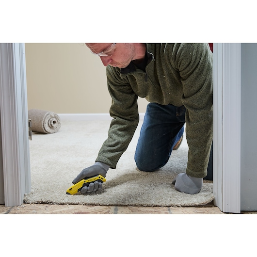 A person wearing work gloves is kneeling on a carpet and using a yellow STANLEY utility knife to cut the edge of the carpet near a doorway. A roll of carpet is visible in the background.