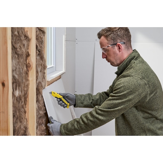 A person wearing gloves uses a yellow STANLEY utility knife to cut a piece of drywall near a window opening inside a building under construction.
