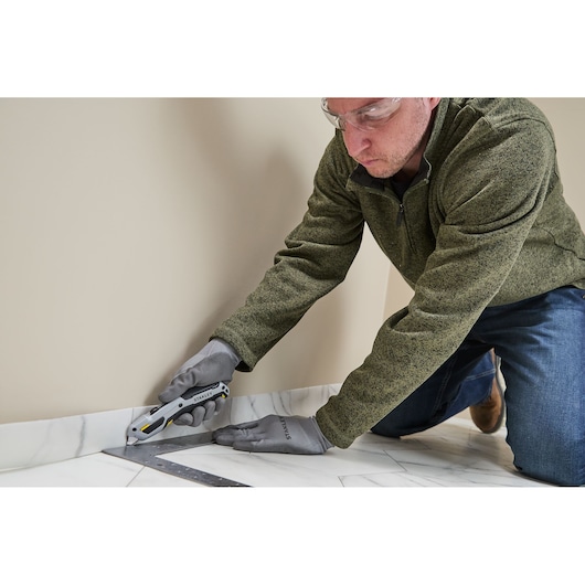 A person wearing gloves and a green jacket is using a STANLEY utility knife (model STHT10721-0) to cut along the edge of a floor near a wall, with a metal square ruler for guidance.