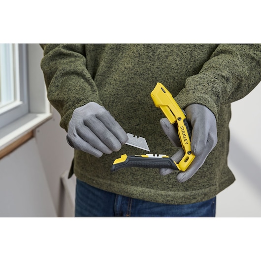 A person wearing gray gloves is holding a STANLEY folding utility knife, inserting or removing a blade. The knife has a yellow and black handle.