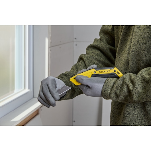 A person wearing grey gloves is holding a yellow STANLEY utility knife and changing its blade indoors near a window.