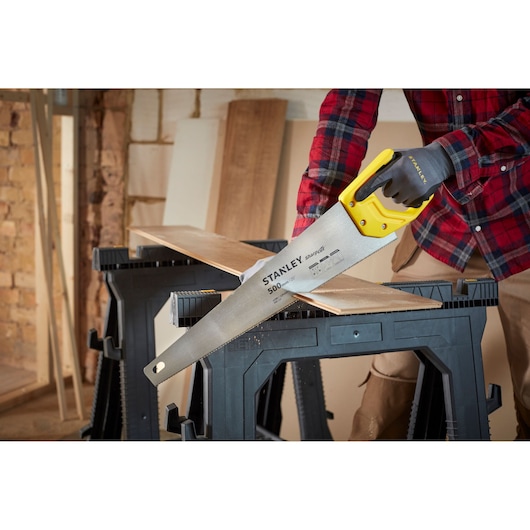 STANLEY STHT20371-1 hand saw with yellow handle cutting wood plank on sawhorse, photographed at an angle.