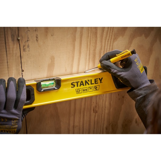 A person wearing work gloves uses a yellow STANLEY spirit level to mark a straight line on a wooden surface.