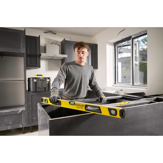 A person in work gloves is using a yellow STANLEY spirit level in a modern kitchen under construction. Cabinets, a toolbox, and a window can be seen in the background.