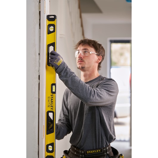 A person wearing work gloves and a tool belt uses a yellow STANLEY spirit level to check the vertical alignment of a door frame inside a building.