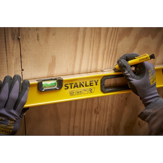 Close-up of gloved hands using a yellow STANLEY spirit level and utility knife to mark a straight line on a wooden board.