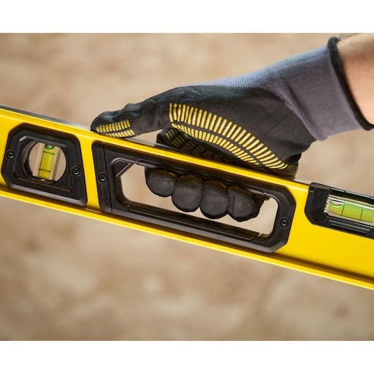 A close-up of a hand wearing a work glove holding a yellow spirit level with built-in handle and visible vials for measuring angles.