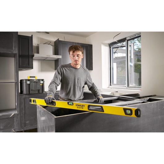 A person with a blurred face is using a STANLEY spirit level (model STHT41048-0) to check alignment during construction or installation in a modern kitchen. The spirit level is yellow with black accents and has visible bubble vials. A toolbox is seen in the background.