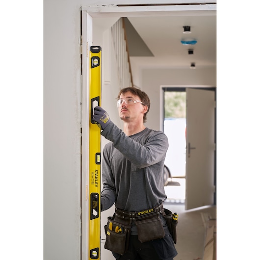 Person using a yellow STANLEY spirit level to measure the vertical alignment of a door frame. The individual is wearing work gloves, a tool belt, and casual work attire in an indoor setting.