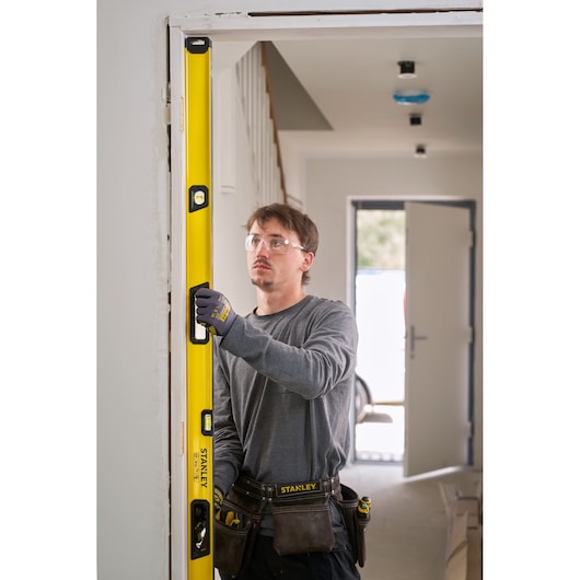 A person wearing work gloves and a tool belt is using a yellow STANLEY spirit level to check the vertical alignment of a door frame in a house under construction or renovation.