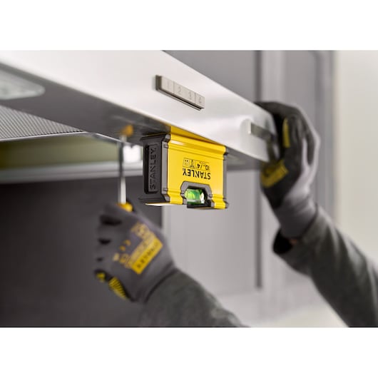 A person wearing gloves uses a yellow STANLEY spirit level (model STHT41704-0) to align a metallic surface, likely during a home improvement or installation task.