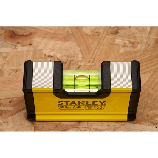 A Stanley STHT41704-0 10cm/4in spirit level tool with a yellow body and black ends, placed on a wooden surface.