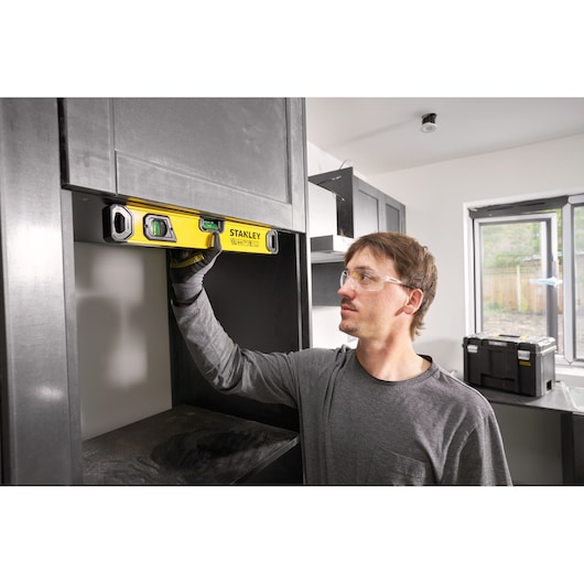 A person using a STANLEY spirit level (model STHT41716-0) to check the alignment of a shelf inside a modern kitchen or workshop. The person's face is blurred for privacy. There are black cabinets and a tool box visible in the background.