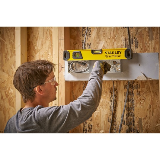 A person using a STANLEY spirit level to check the alignment of an electrical box on a wooden wall during construction. The person is wearing a grey shirt and work gloves.