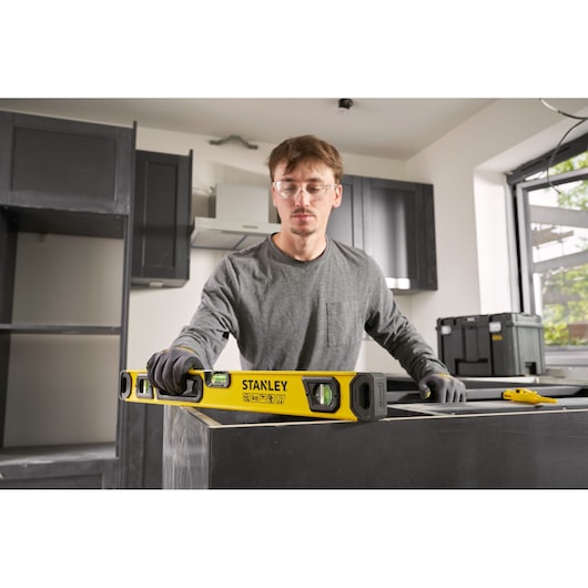 A person wearing gloves is using a STANLEY spirit level (model STHT41724-0) to check the level of a surface in a kitchen under construction. Other tools and storage boxes are visible in the background.