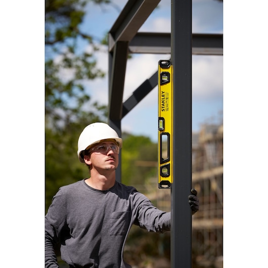 A person wearing a white safety helmet uses a yellow STANLEY spirit level to check the alignment of a vertical metal beam at a construction site. The person's face is blurred.