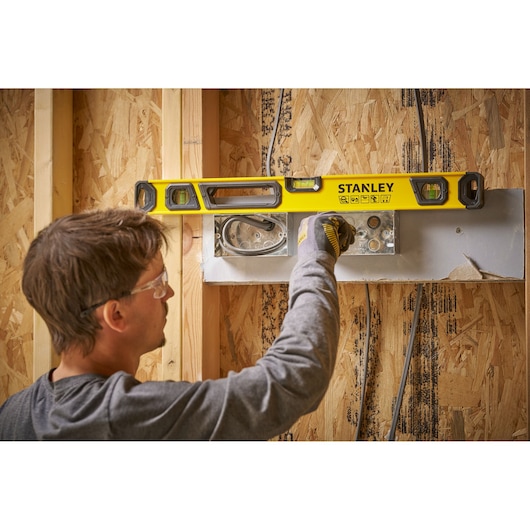 A person using a yellow STANLEY spirit level to check the alignment of an installation on a wooden wall under construction.