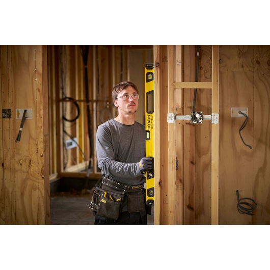 A person with their face blurred is holding a yellow STANLEY spirit level against a wooden wall frame on a construction site. The person is wearing work gloves and a tool belt with various tools.