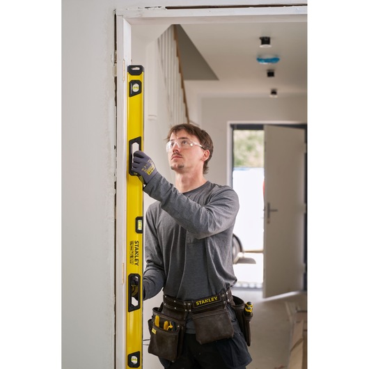 A person wearing work gloves and a tool belt is using a yellow STANLEY spirit level to check the vertical alignment of a door frame in an indoor construction setting. The person's face is blurred.