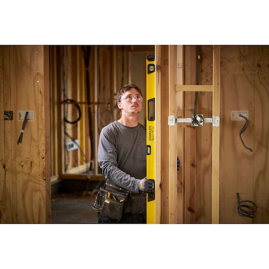 A person wearing work gloves and a tool belt uses a yellow STANLEY spirit level to check the vertical alignment of a wooden stud frame in a construction site. The background shows unfinished wooden walls and electrical wiring.