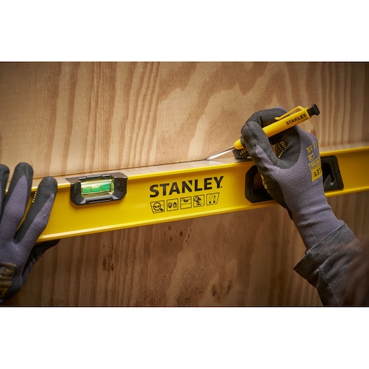 A person wearing gloves uses a STANLEY yellow spirit level and a pencil to mark a straight line on a wooden surface.