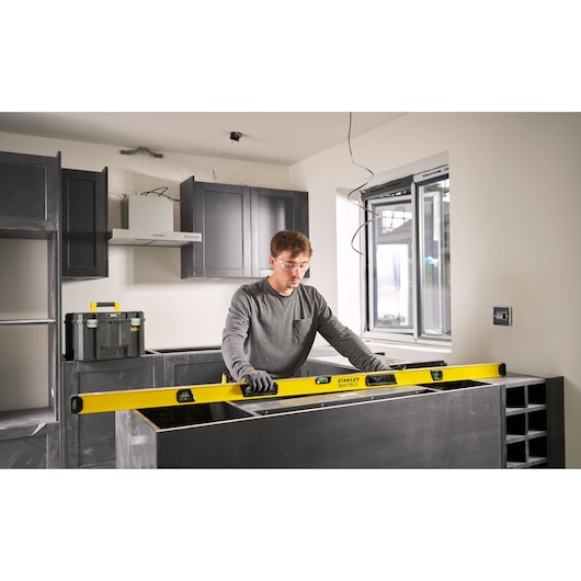 A person using a STANLEY spirit level (model STHT41772-0) to check the alignment of a kitchen countertop during installation. The person is wearing gloves and is working in a modern kitchen under construction.