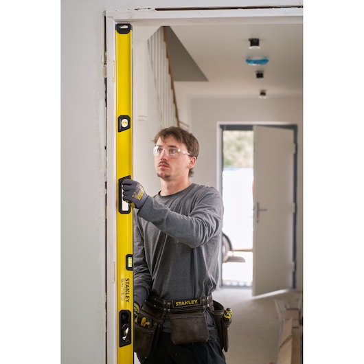 A person wearing work gloves and a tool belt is using a yellow STANLEY spirit level to check the vertical alignment of a door frame in an indoor setting.