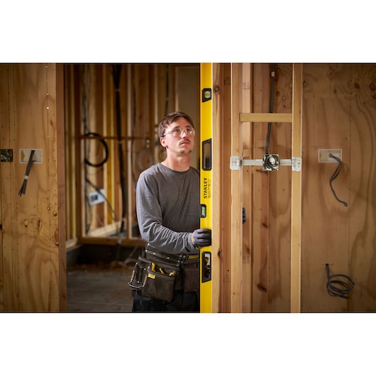 A person with a blurred face using a yellow STANLEY spirit level to check the vertical alignment of wooden framing at a construction site. The individual is wearing work gloves and a tool belt, and the surroundings show unfinished wooden walls and electrical wiring.
