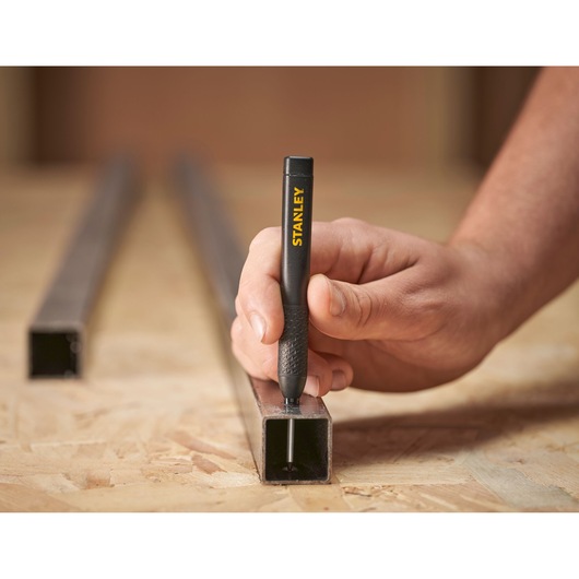 Hand using a Stanley marking pen to write on a metal tube placed on a wooden work surface.