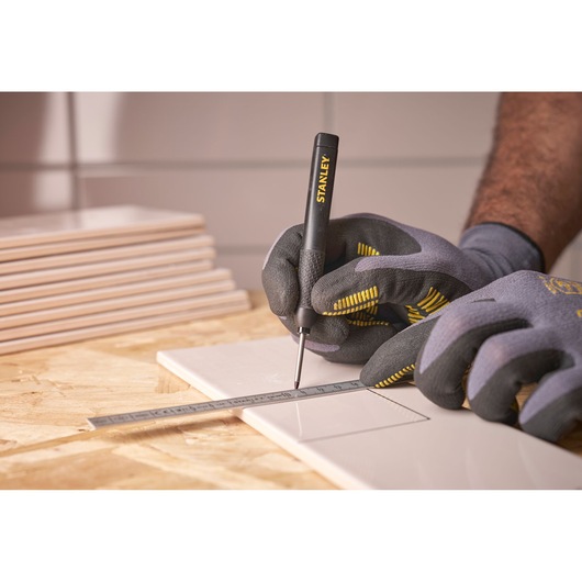 Gloved hands use a STANLEY marking tool and ruler to draw lines on a tile for precise measurement.