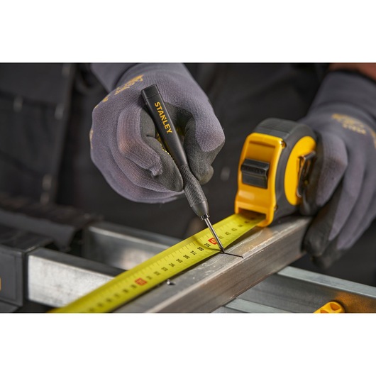 Gloved hands use a Stanley tool and tape measure to mark a metal beam for precise measurement.