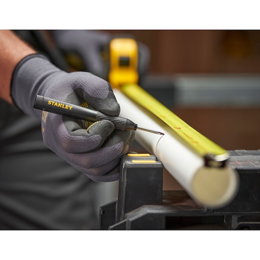 Gloved hand uses a STANLEY marking tool and tape measure to mark a white pipe in a workshop setting.