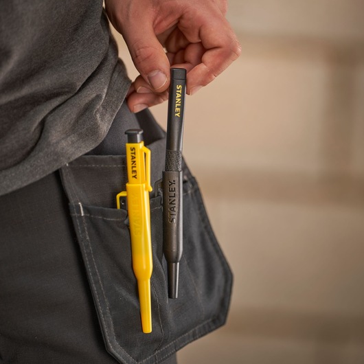 Stanley marking tools in a pocket, one yellow and one black, held by a hand, worn on work trousers.